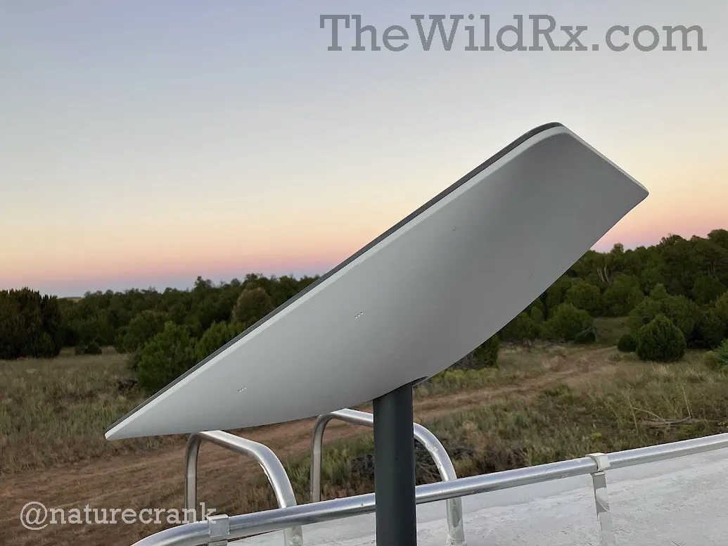 A Starlink satellite dish mounted on top of an RV parked in a northern Arizona scrub forest with juniper trees and a clear sky.