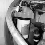 A close-up, black and white view of an RV water pump assembly under a sink, showing a Seaflo professional grade pump connected to reinforced flexible water lines and PEX tubing.