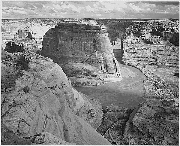 Black and white landscape photography by Ansel Adams showing the sheer sandstone cliffs and winding river of Canyon de Chelly National Monument in Arizona.