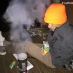 A hiker in an orange beanie sits on a wooden porch using a portable stove to cook a meal during a multi-day backpacking trip.