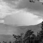 A black and white mountain landscape showing a heavy rain shaft and virga falling from dark clouds over a forested valley.