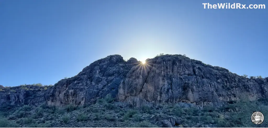 A sunburst effect created by the sun peeking over the sharp summit of a rocky desert mountain peak in a clear blue sky.