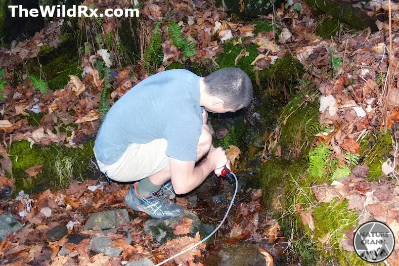 A hiker crouching next to a small forest stream on the Appalachian Trail using a manual pump water filter.