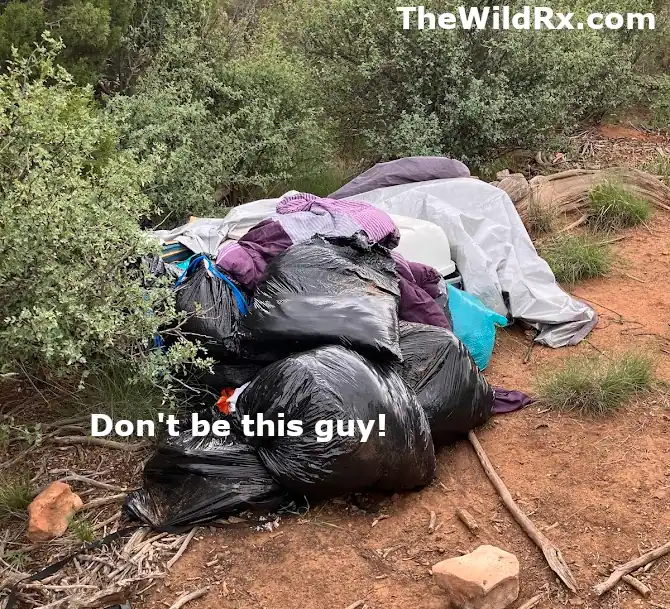 A pile of black trash bags and abandoned camping gear left behind in a natural desert environment with brush and red dirt.