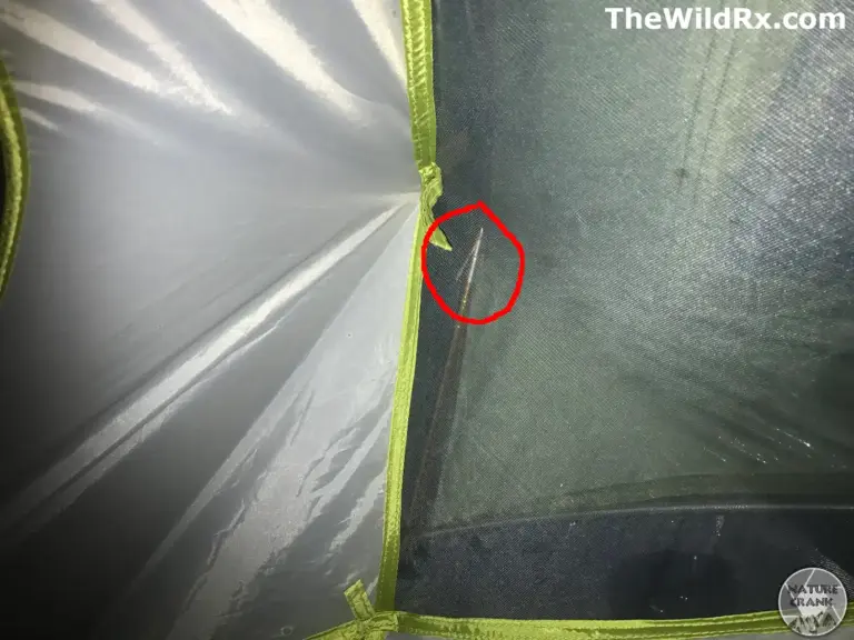 A close-up view from inside a tent showing a broken orange aluminum tent pole with a sharp, jagged fracture visible through the green mosquito mesh. The break is highlighted with a red circle.