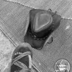 A high-angle black and white shot of a cowboy hat and a hiker's foot in a Chaco sandal with socks on a textured groundsheet, representing the minimalist cowboy camping lifestyle.