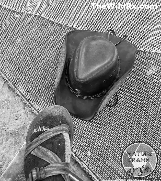 A high-angle black and white shot of a cowboy hat and a hiker's foot in a Chaco sandal with socks on a textured groundsheet, representing the minimalist cowboy camping lifestyle.
