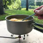 A hand stirring shrimp and pasta in a titanium backpacking pot on a remote canister pocket stove during a gear test.