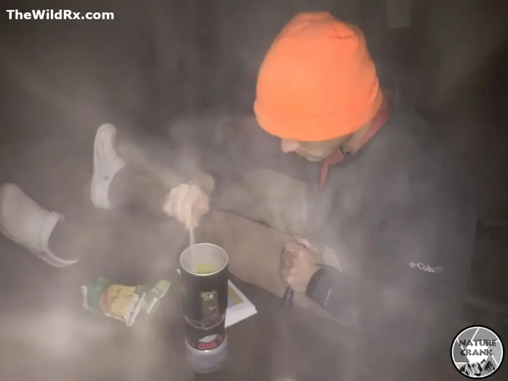 A hiker (my friend) in an orange beanie and Columbia jacket stirring a steaming pot of food on a camp stove during a winter backpacking trip.