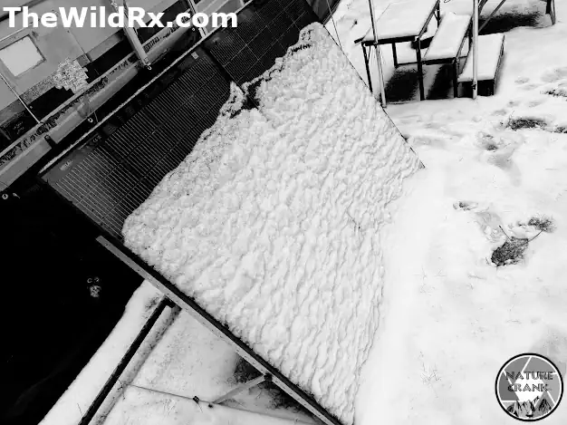 A portable ground-mounted RV solar panel array partially covered in thick snow next to a camper during winter boondocking.